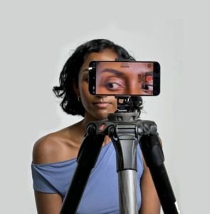 Shakti Mb holding a phone on a tripod in front of her face, the screen showing her eyes